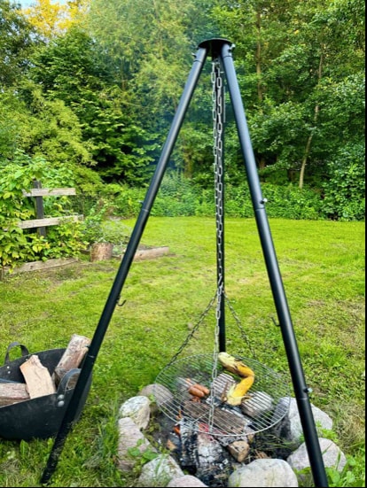 A fire pit setup features a black tripod frame with a cooking grill suspended over the embers. Nearby, a basket holds firewood. The grassy surroundings include dense greenery, contributing to a natural and inviting outdoor cooking space.