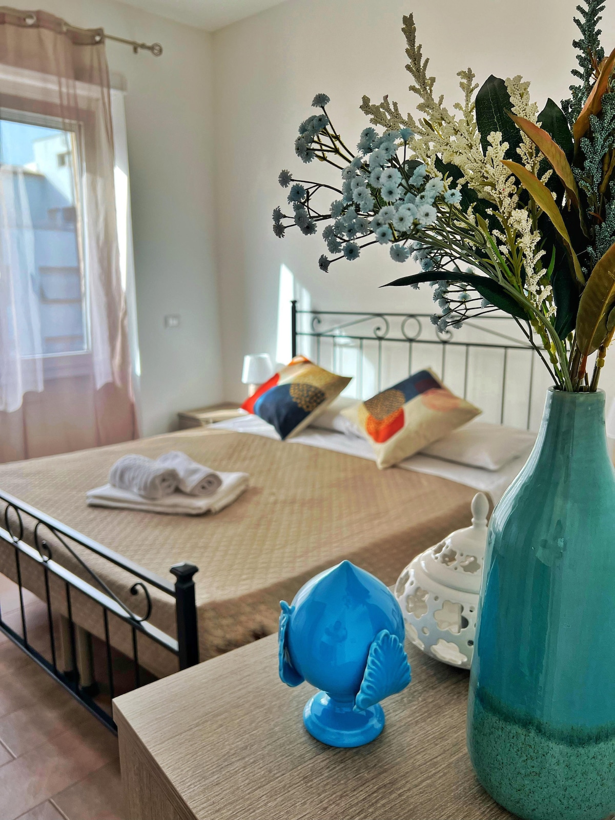 A well-lit bedroom features a spacious bed adorned with decorative pillows and neatly arranged towels. A bedside table holds a vibrant blue vase filled with faux flowers, complementing the room's calming decor. Natural light streams in through the window, enhancing the inviting atmosphere.