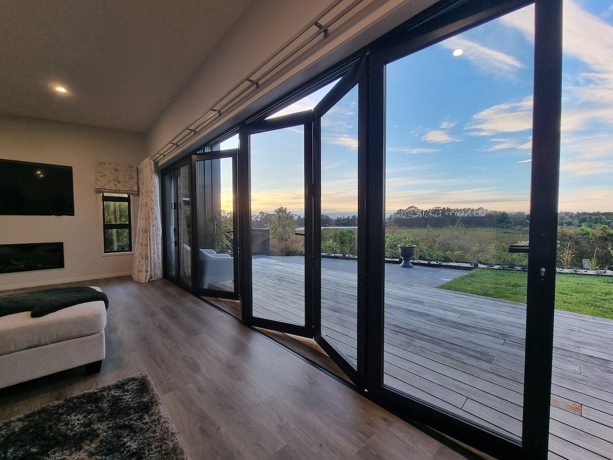 Large bifold doors open to reveal an expansive outdoor deck, providing views of lush greenery and a clear sky. The interior space is bright, with wooden flooring and a cozy seating area visible. A subtle gas fire is positioned against the far wall.