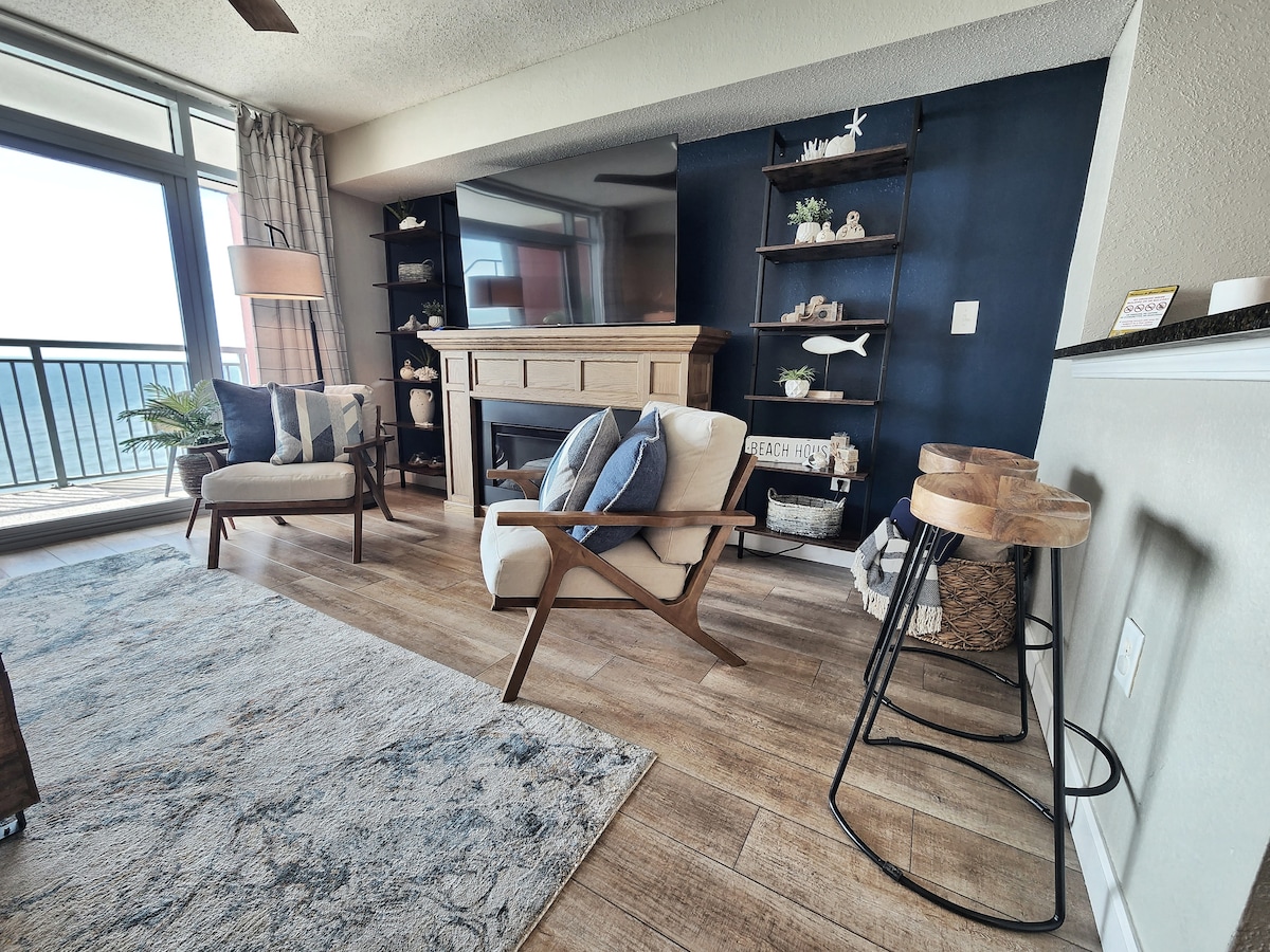 The living area features a neutral color palette with a textured area rug and hardwood flooring. A wooden fireplace surrounds a large television, while a wall shelf displays decorative items. Seating includes two stylish chairs and two wooden bar stools, with a view of the ocean visible through floor-to-ceiling windows.