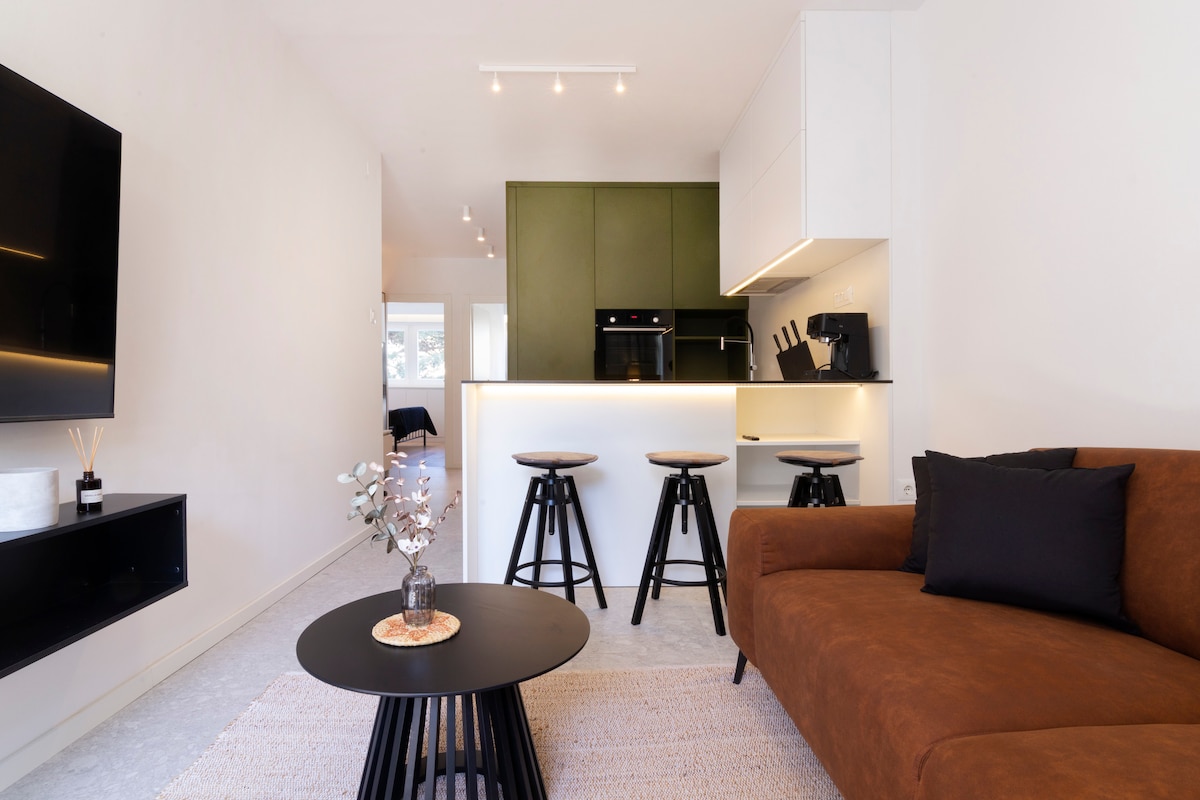 A modern living area features a brown sectional sofa and a round black coffee table adorned with a decorative centerpiece. Three black stools are positioned around a sleek kitchen island with olive green cabinetry, enhancing the open layout. Light fixtures above provide even illumination.