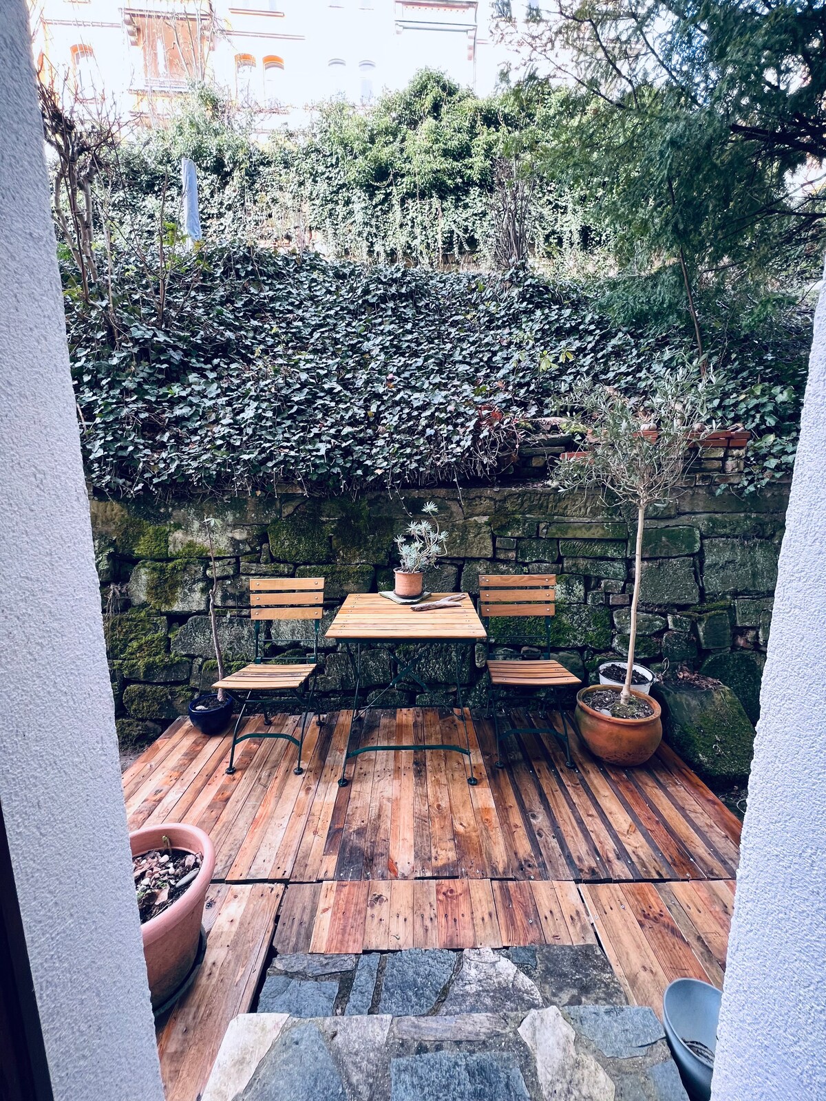 A small private terrace is shown, featuring a wooden table with two matching chairs. Potted plants are placed around the area, and textured stone walls provide a natural backdrop. The space is surrounded by greenery, creating a quiet spot for relaxation.