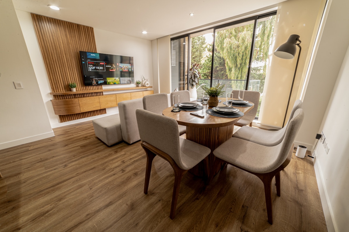 A dining area showcases a round wooden table surrounded by six upholstered chairs. A light fixture stands beside the table, and a media console with a large TV is mounted on the wall. Natural light enters through a glass door leading to an outdoor space.