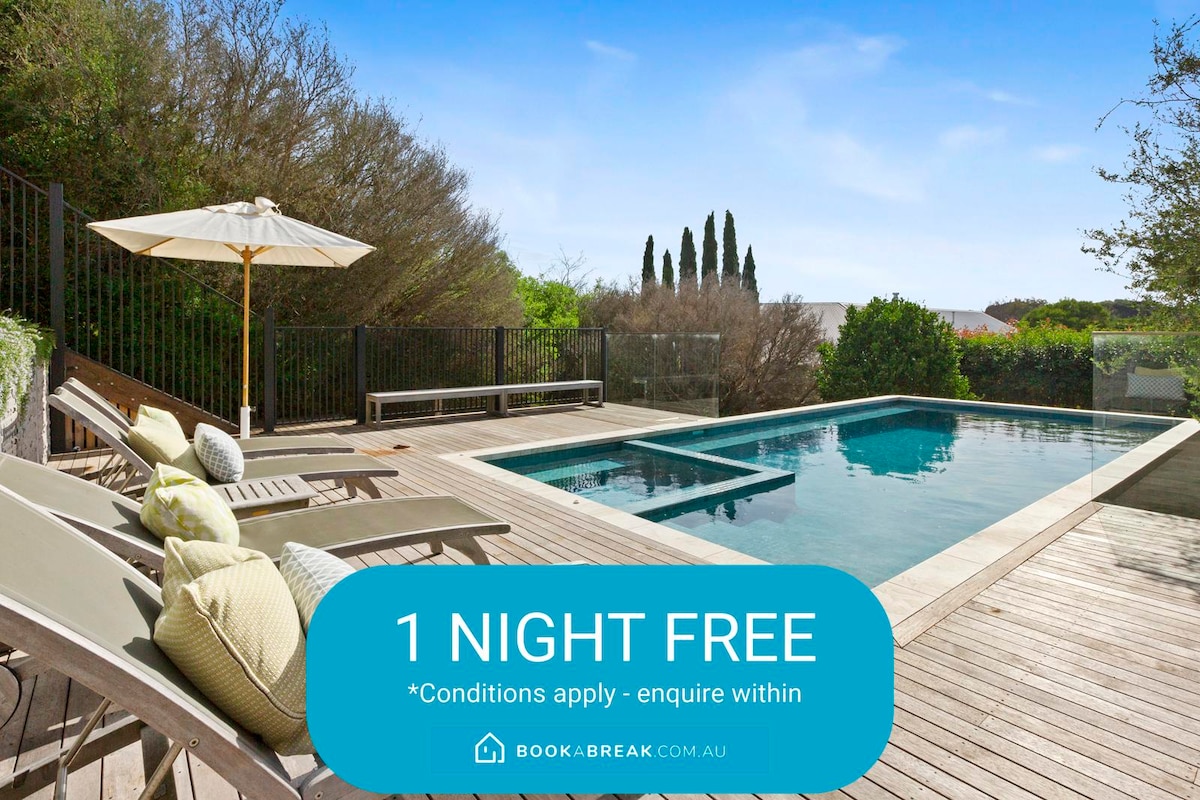 A serene outdoor space is presented, featuring a gas-heated pool surrounded by a sun terrace with lounge chairs. A large umbrella provides shade alongside the pool. Lush greenery borders the area, enhancing privacy and contributing to a tranquil atmosphere.