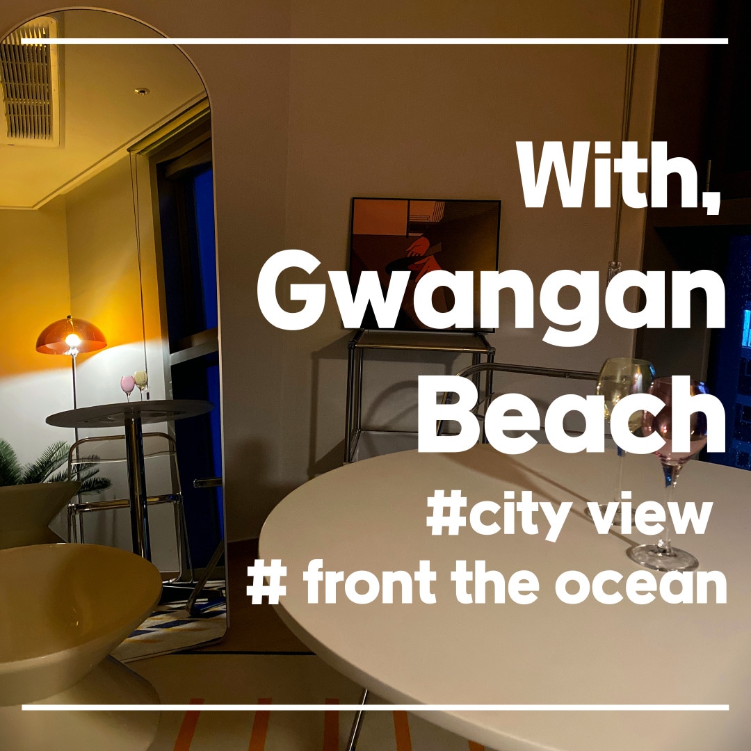 The studio features a modern table with two glasses set atop it, positioned near a large mirror. A gentle glow from a stylish lamp illuminates the space, highlighting the inviting city view and the nearby ocean through expansive windows.