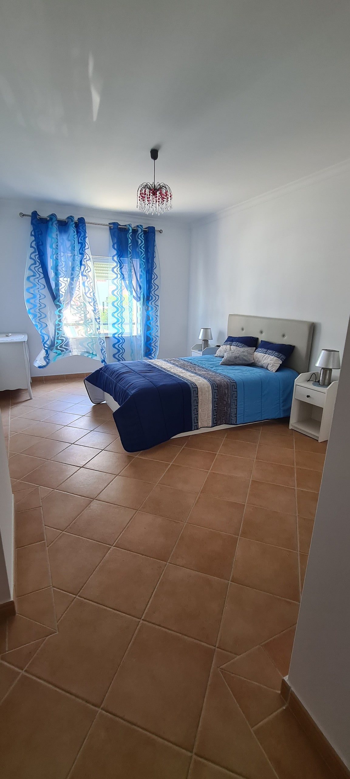 A well-lit bedroom is depicted with a spacious bed covered in blue and grey bedding. Decorative curtains frame a window that allows natural light to fill the room. Two bedside tables with lamps are placed on either side of the bed, offering a comfortable layout.