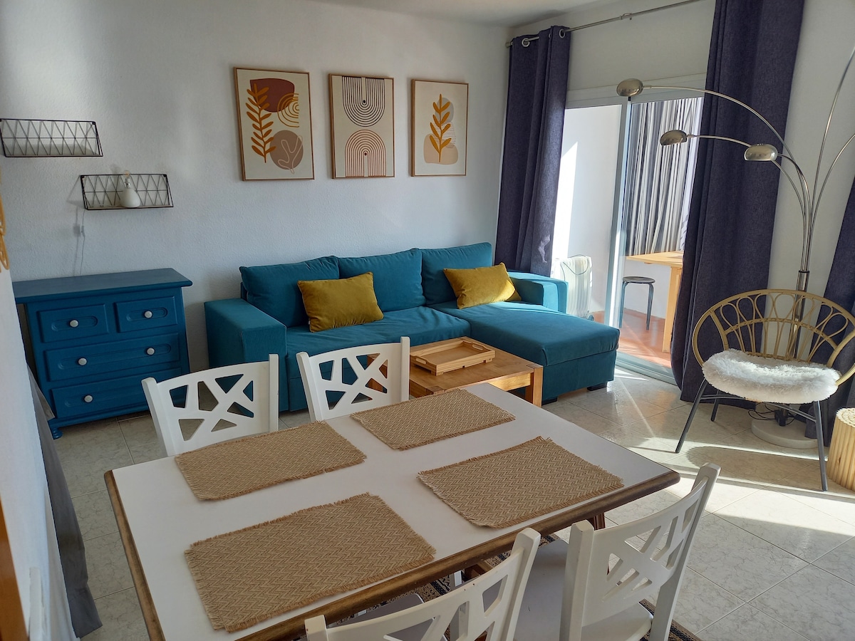 A bright and inviting living space features a blue sofa with two yellow cushions, accompanied by a light wooden coffee table. A dining table with six chairs sits nearby, showcasing woven placemats. Large window curtains frame natural light entering from the balcony.