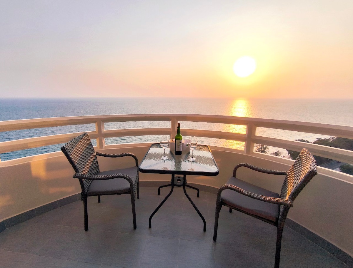 A private balcony offers a stunning view of the sea at sunset. A glass-top table is set with two glasses and a bottle, accompanied by two woven chairs. The soft glow of the sun reflects off the water, creating a serene atmosphere.