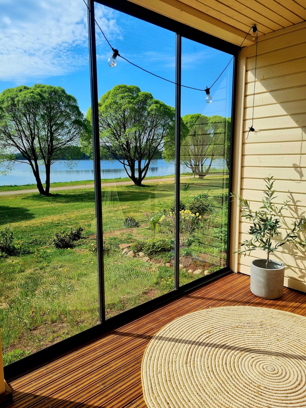 A glass-enclosed terrace offers views of a serene landscape featuring lush green trees and a calm body of water. A round, textured rug lies on the wooden floor, and a potted plant adds a touch of nature to the interior space.