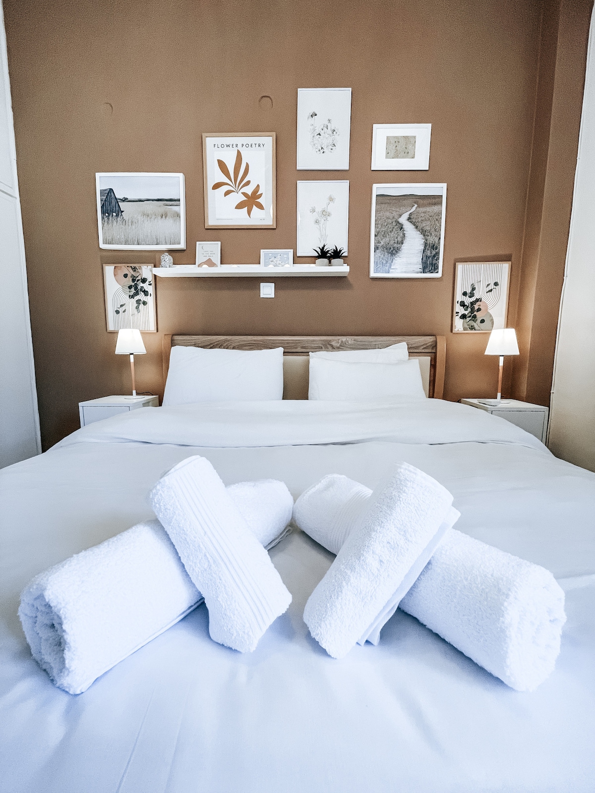 A warm-toned bedroom is presented, featuring a spacious bed with soft linens. Four neatly rolled white towels are arranged in the foreground. Framed artwork decorates the walls, complemented by two bedside lamps providing gentle lighting.