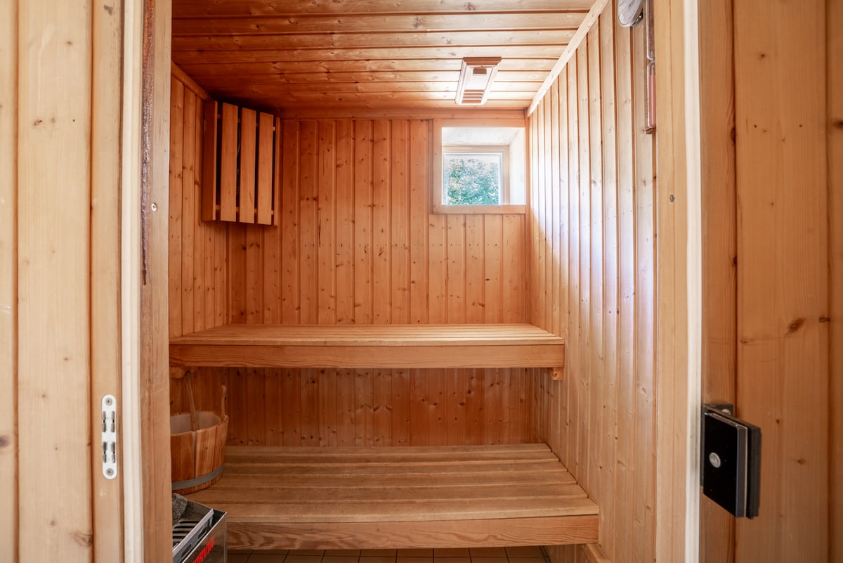 A wooden sauna is presented with smooth paneling and warm tones, featuring two benches arranged on either side. A small window allows natural light to enter, while a wooden bucket is placed against one wall, enhancing the soothing environment.