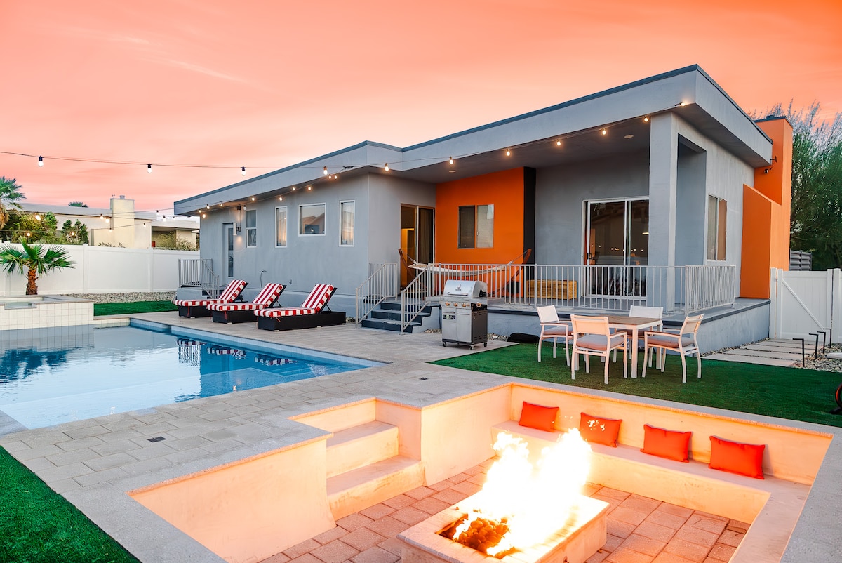 The exterior of the home features a modern design with bold orange accents and large windows. A pool glistens beside the patio, which includes a seating area and a barbecue grill. A fire pit with orange cushions is visible, surrounded by lounge chairs in a relaxing outdoor setting.