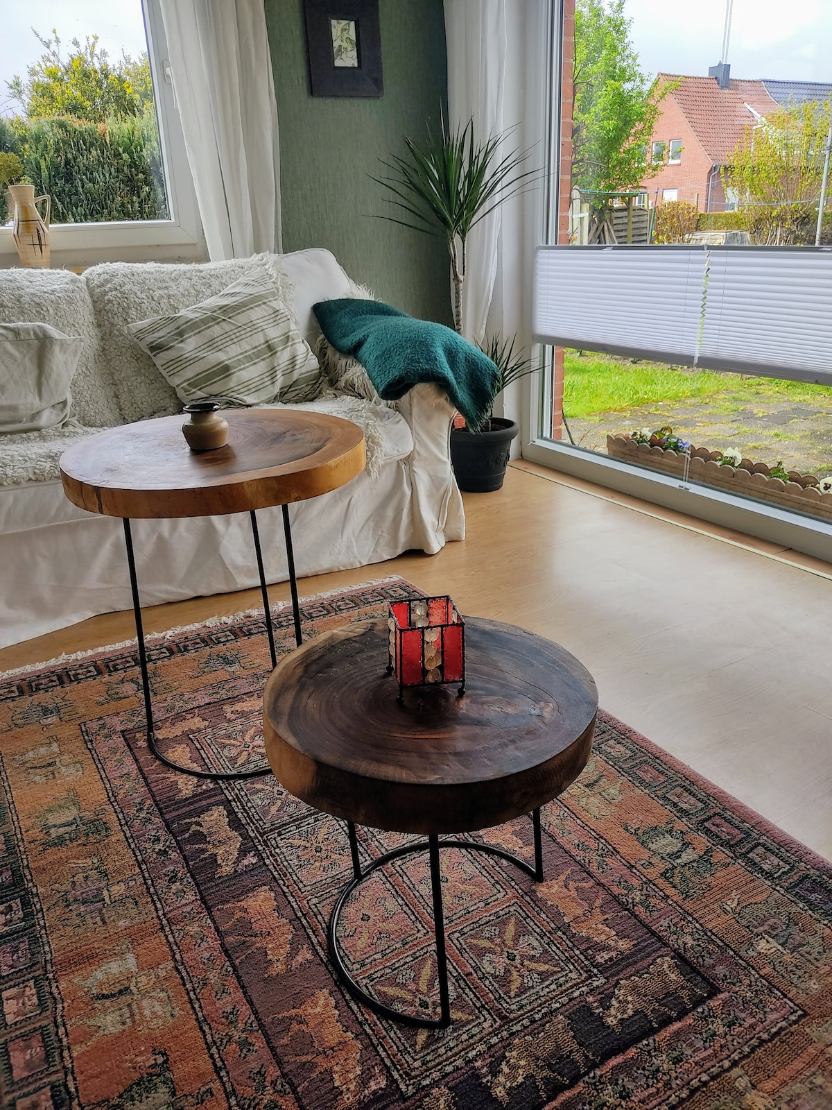 Two wooden side tables are positioned beside a light-colored sofa, adorned with a textured throw. A small red lantern rests on one table, while a plant sits in a pot nearby. Natural light streams through a large window, illuminating the space.