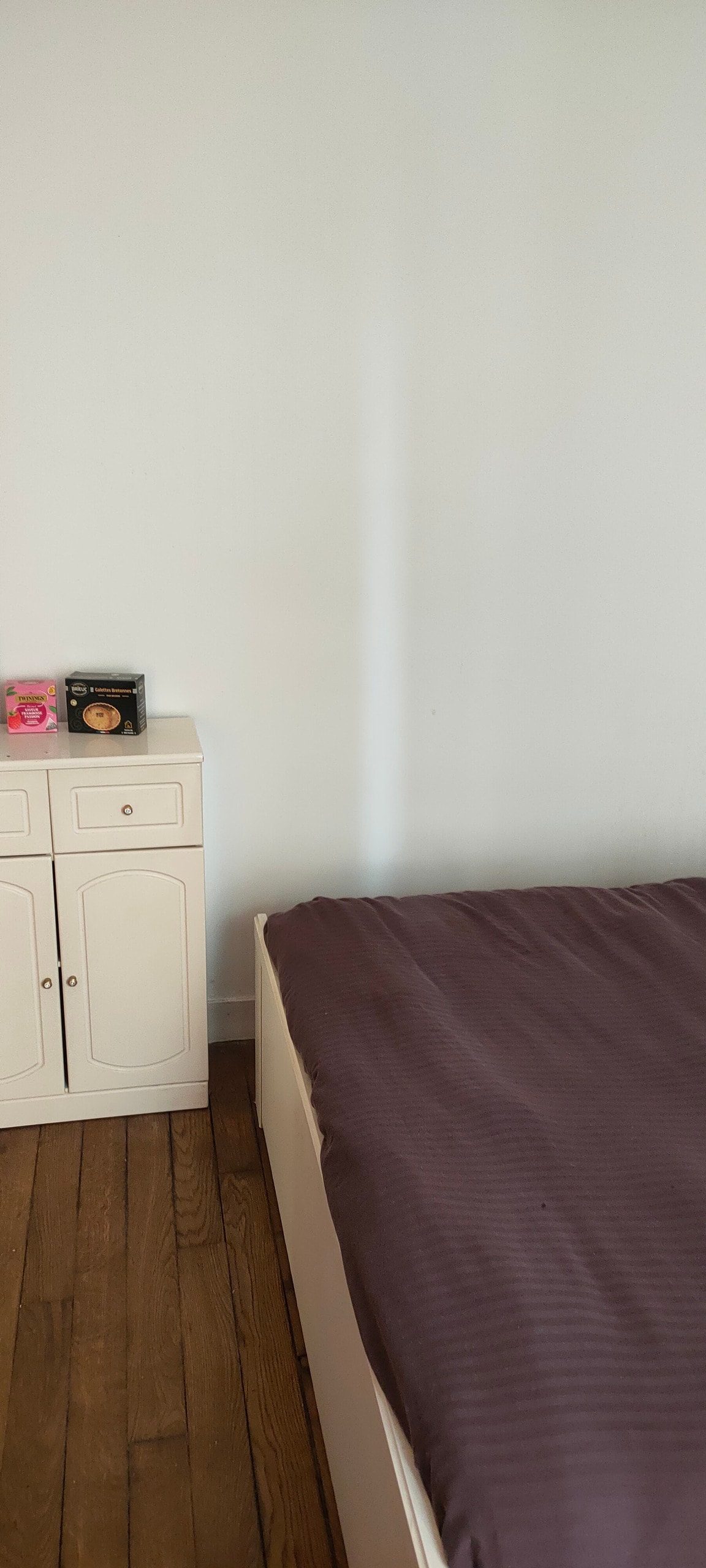 A spacious bed is positioned alongside a light wood cabinet. The cabinet features two door compartments and is adorned with a few decorative items. The wall behind is painted white, accentuating the wooden floor.