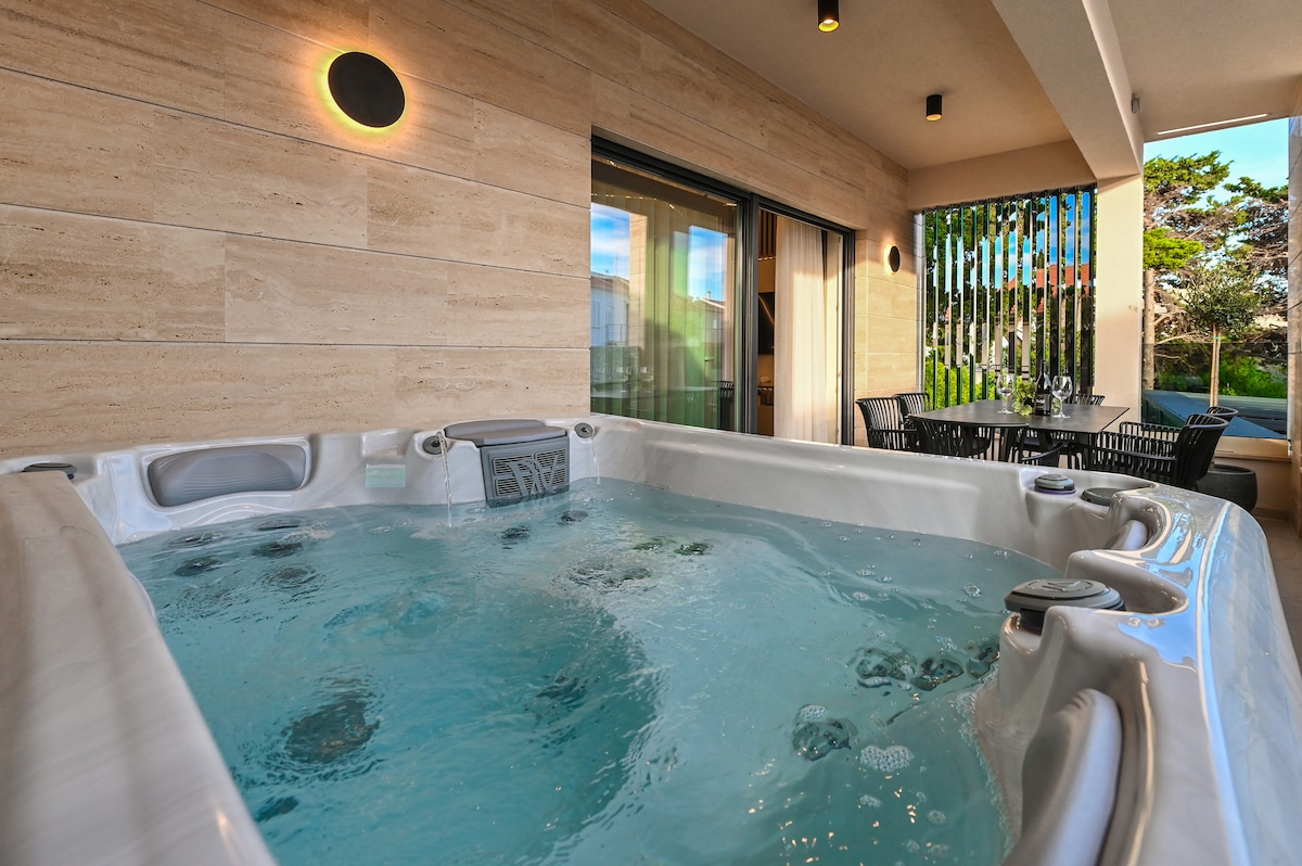 A spacious terrace is showcased, featuring a jacuzzi with bubbling water. An outdoor dining area with a table and chairs is visible nearby, surrounded by greenery. The area is illuminated by soft lighting, creating a relaxing atmosphere for unwinding.