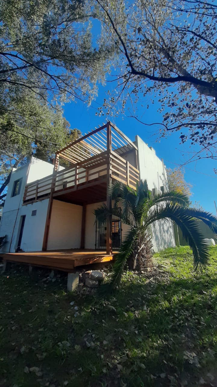 The exterior view of the building highlights a wooden terrace with railings, surrounded by lush greenery and trees. The structure features a white façade, and sunlight illuminates the outdoor space, creating a warm atmosphere.