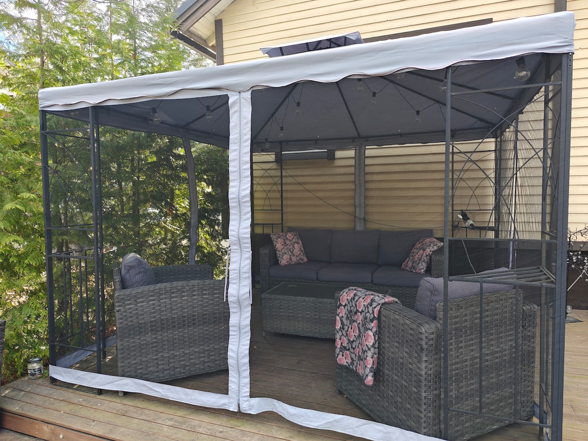 An outdoor seating area is enclosed within a canopy, featuring comfortable wicker furniture. Soft cushions add to the inviting space, which includes a sectional sofa and a coffee table. The surrounding greenery enhances the setting, creating a serene atmosphere for relaxation.