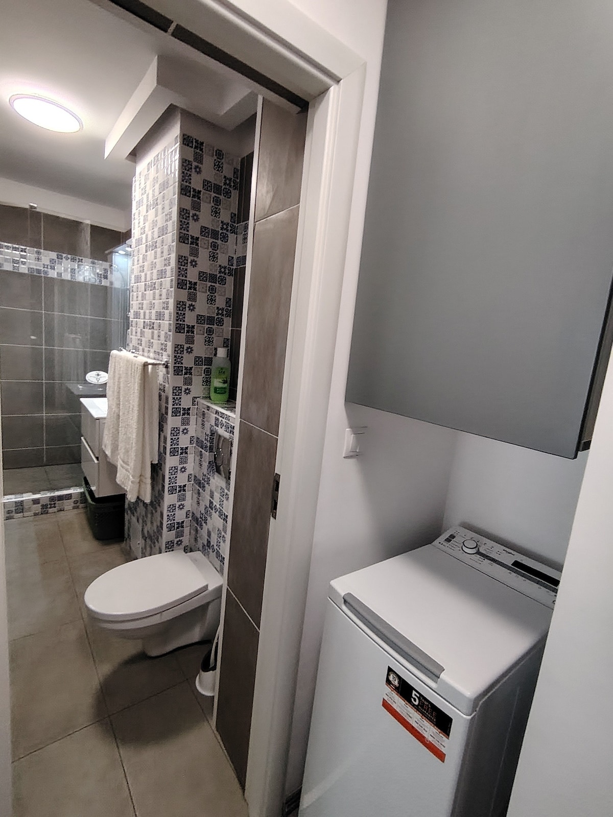 A modern bathroom is captured, featuring a mix of patterned tiles and sleek gray walls. A compact laundry area is visible beside a toilet. A light fixture above the space adds illumination, while a towel hangs neatly on the wall.