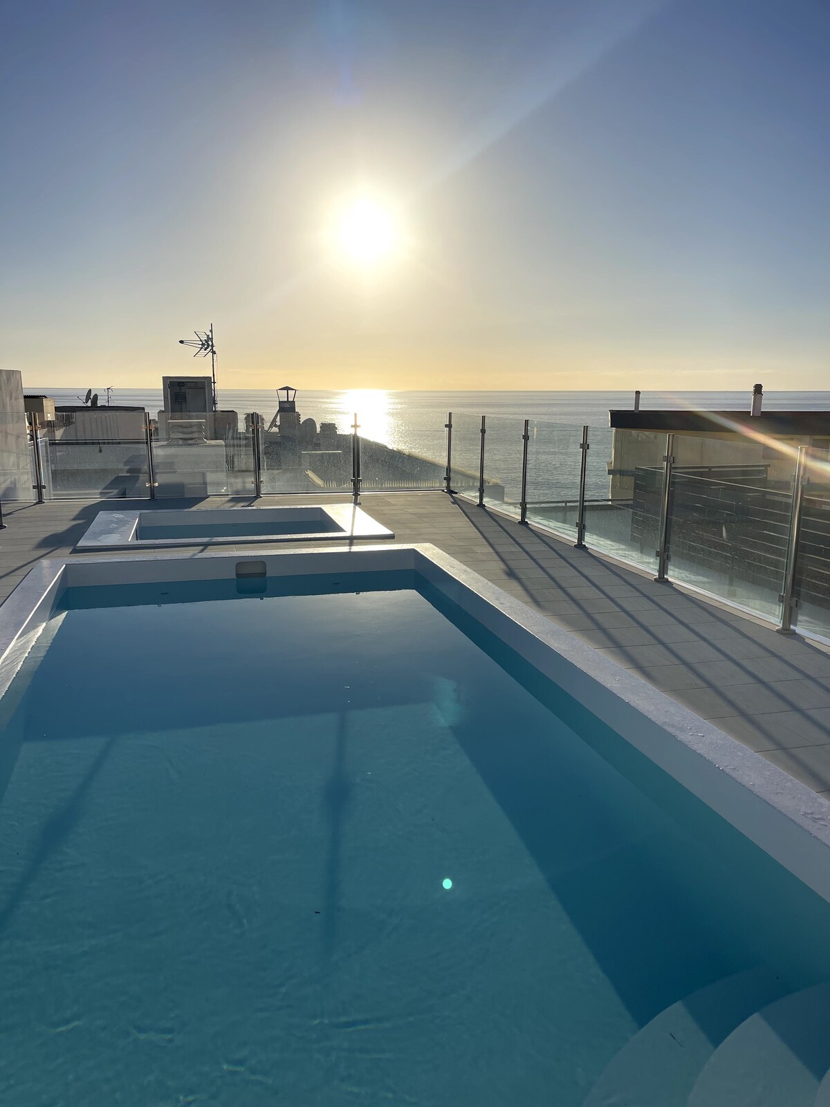 A rooftop area features a small pool bordered by glass railings, reflecting sunlight. The serene sea extends in the distance under a clear sky, and the sun sets on the horizon, creating a tranquil ambiance.