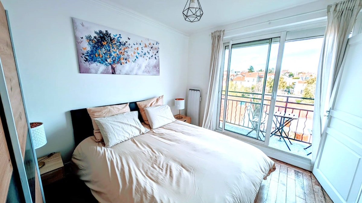 A bright bedroom features a spacious bed with neutral bedding, complemented by bedside lamps. A sliding glass door opens to a small balcony with a table and chairs, providing an outdoor view filled with greenery. Natural light enhances the calming atmosphere.
