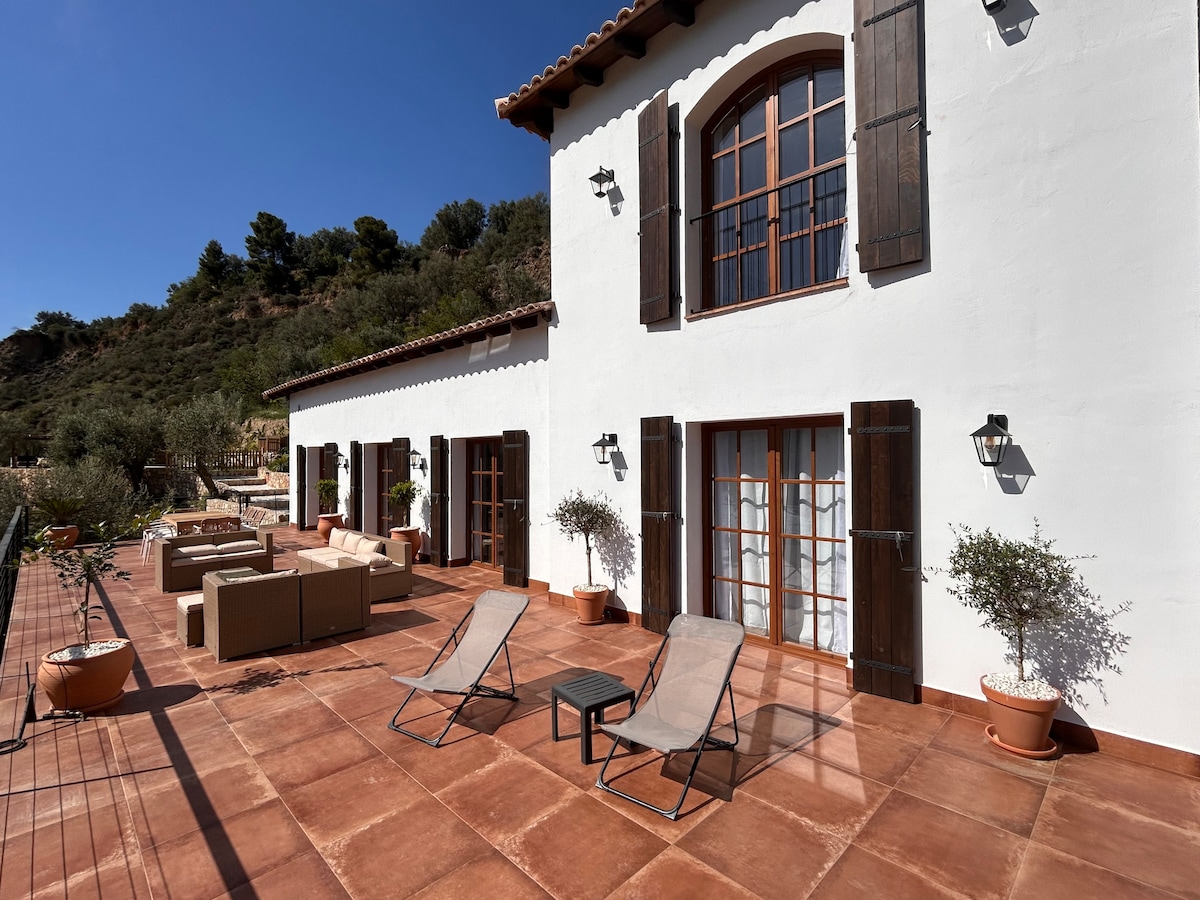The exterior terrace features a spacious layout with two lounge chairs and a small table, complemented by potted plants. A comfortable seating area is arranged against the wall, with large glass doors leading to the interior. Sunlight enhances the texture of the terracotta tiles.