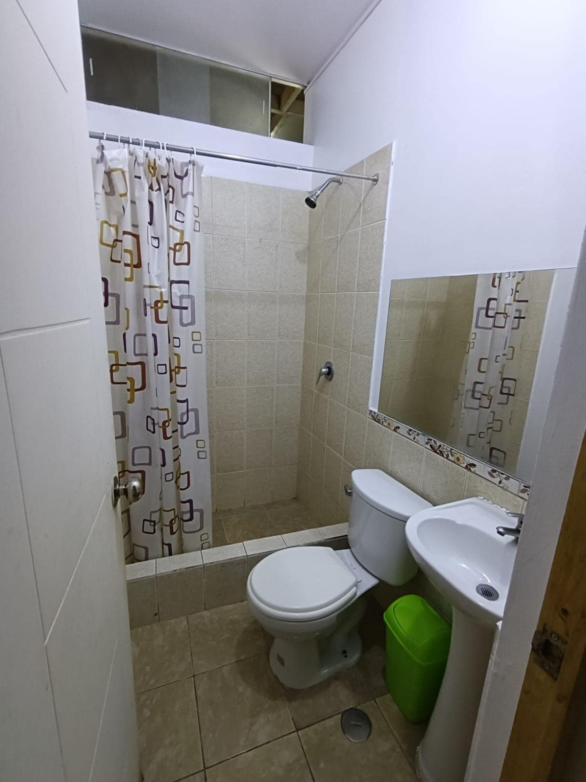 The private bathroom features neutral ceramic tiles and a shower area enclosed by a patterned curtain. A mirror above the sink complements a toilet and a small green trash bin, creating a functional and tidy space.