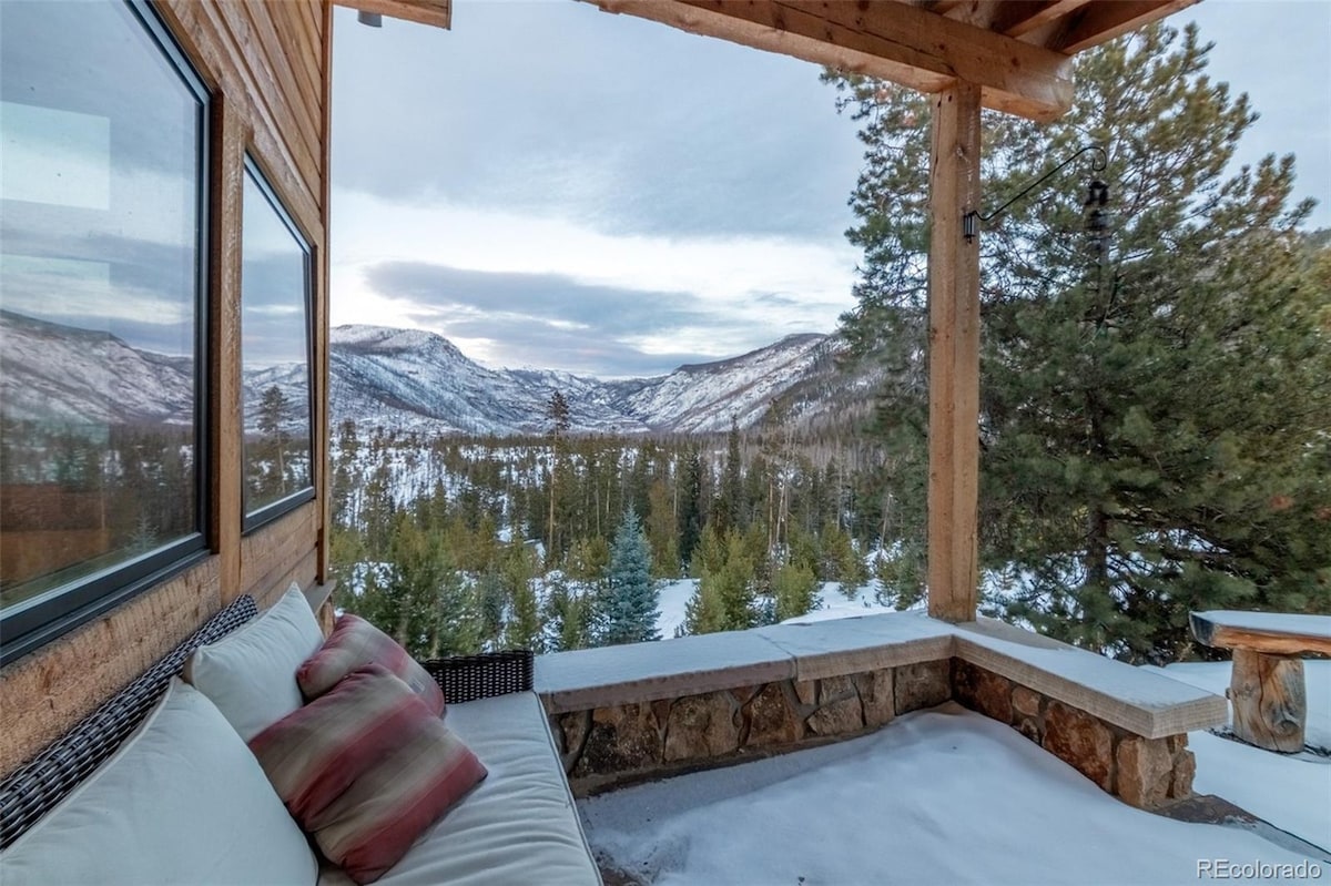 A covered outdoor seating area features a cozy arrangement of cushions against a wooden frame, offering unobstructed views of snow-covered mountains and pine trees. The serene landscape stretches into the distance, enhancing the inviting atmosphere of the space.