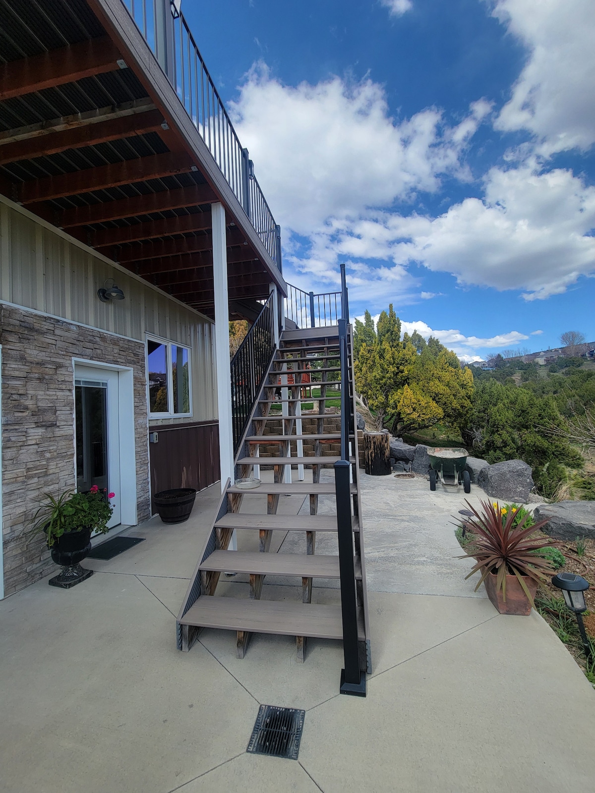 A set of wooden stairs with a modern railing leads up from a paved area to the entrance of a loft space. Lush greenery and decorative plants are visible along the side, complementing the natural surroundings.
