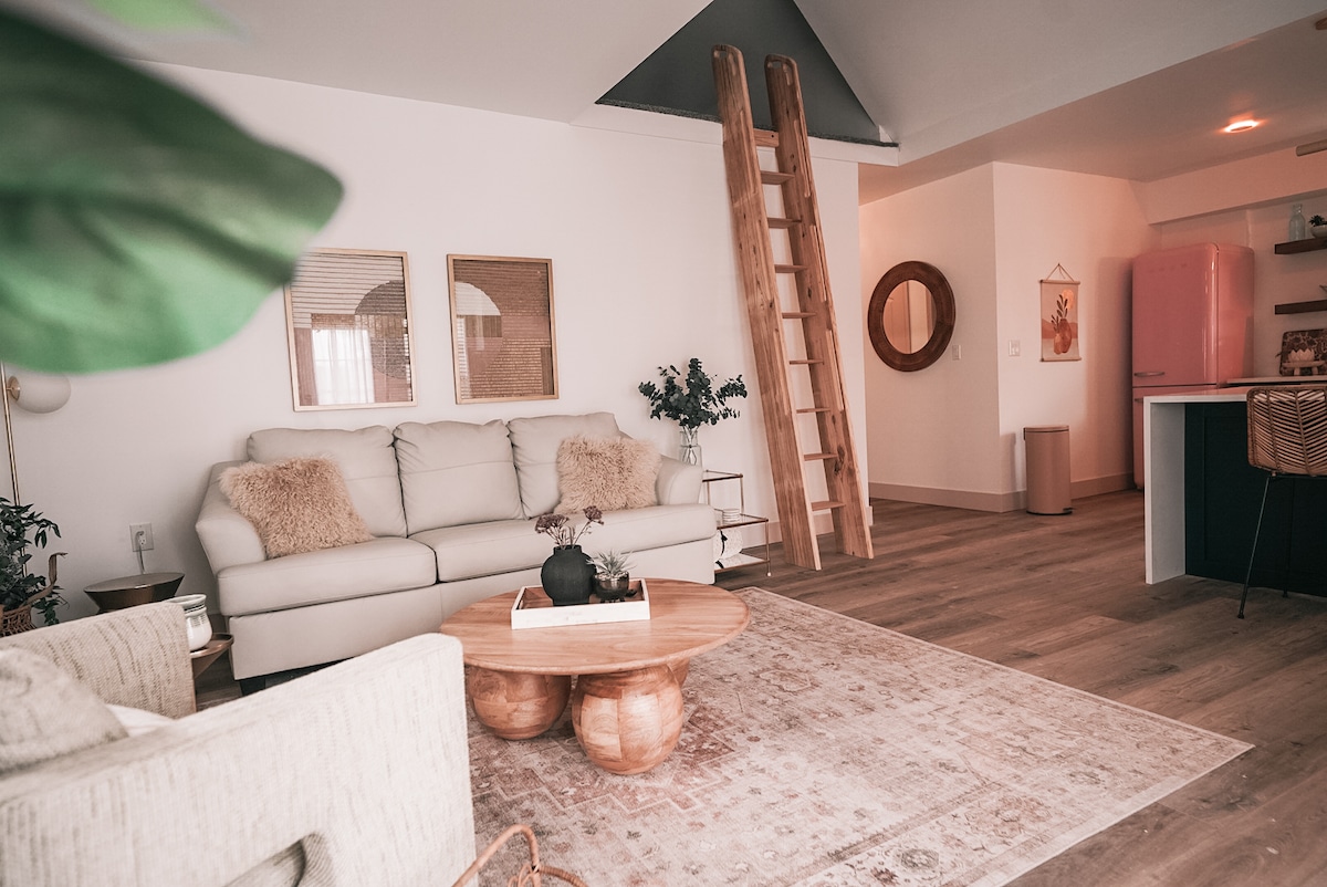 A spacious living area features a light-colored sofa and a round coffee table with a natural wood finish. A decorative rug lies beneath the table, while a ladder leads to a loft space. The pink refrigerator adds a vibrant touch to the modern decor.