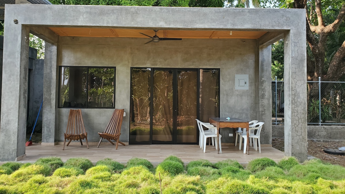 A modern outdoor space features a wooden dining table surrounded by white chairs. Two wooden rocking chairs sit to the left of the table. Large glass doors offer views of the exterior greenery, while a ceiling fan is positioned above the seating area.