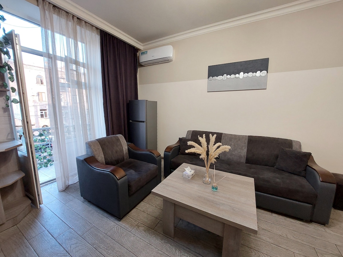 A modern living area is furnished with a comfortable sofa and armchair, positioned around a wooden coffee table. Large windows with sheer curtains allow natural light to fill the space. A sleek refrigerator is visible in the corner, contributing to the functional design.