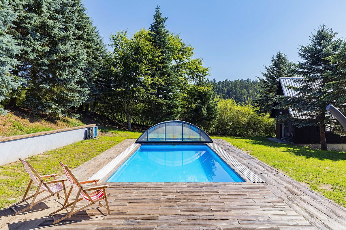 A swimming pool with a clear cover is surrounded by wooden decking, offering a serene setting. Expansive green grass and trees frame the area, contributing to a peaceful outdoor space. Two wooden lounge chairs are positioned adjacent to the pool, inviting relaxation.