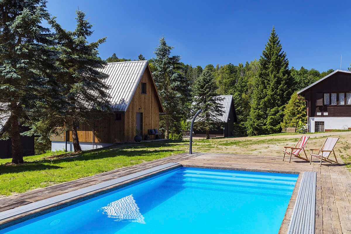 A clear swimming pool is surrounded by a wooden deck and green grass, with two lounge chairs positioned nearby. The scene features a backdrop of tall trees and two modern wooden chalets, blending harmoniously with the natural landscape.