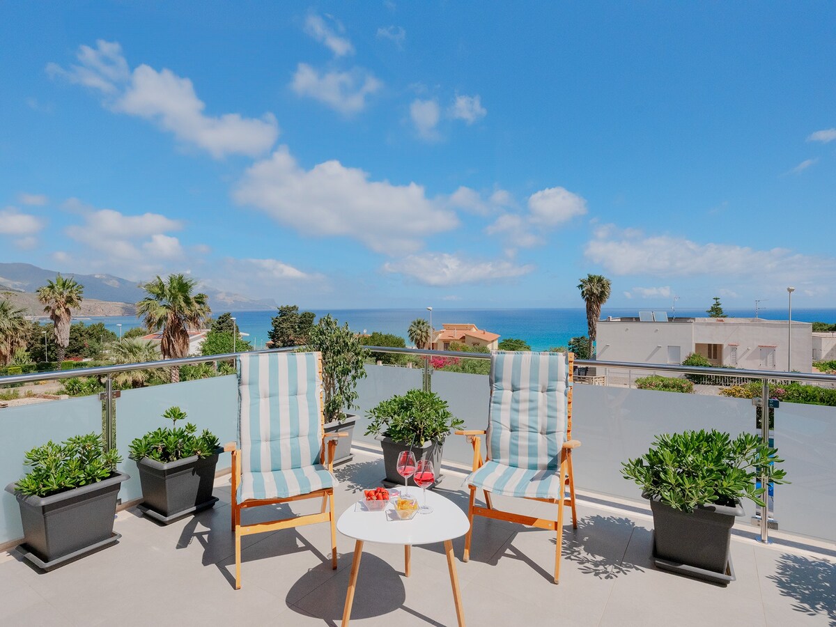 A spacious veranda features two wooden chairs with striped cushions, positioned beside a round table. Potted plants enhance the scene, while a panoramic view of the sea and palm trees is visible in the background under a clear blue sky.