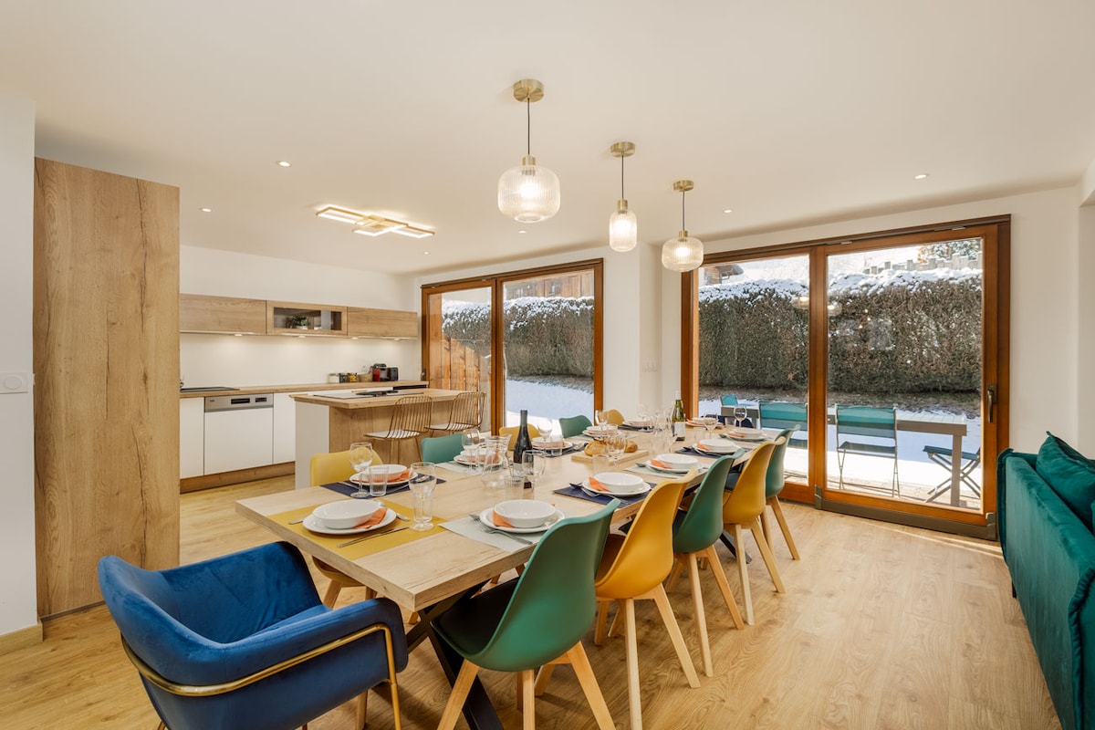 A spacious dining area is highlighted by a large wooden table surrounded by colorful chairs. Large glass doors offer views of the outdoor space, allowing natural light to fill the room. A modern kitchen with sleek cabinetry and appliances is visible in the background.
