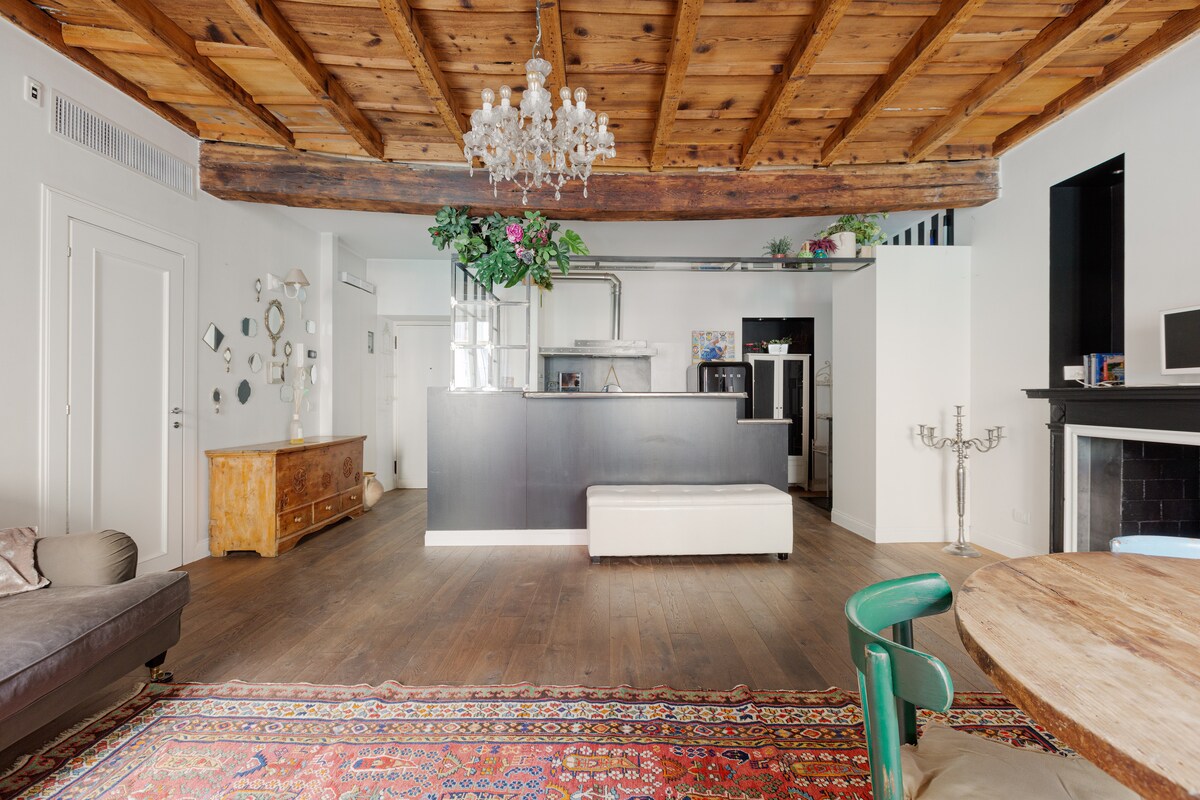 An open living space is highlighted with wooden beams and a chandelier. A modern kitchen area includes a sleek countertop, while a cozy seating area features a dark sofa. A traditional patterned rug covers the floor, complementing the wooden dining table and chairs.