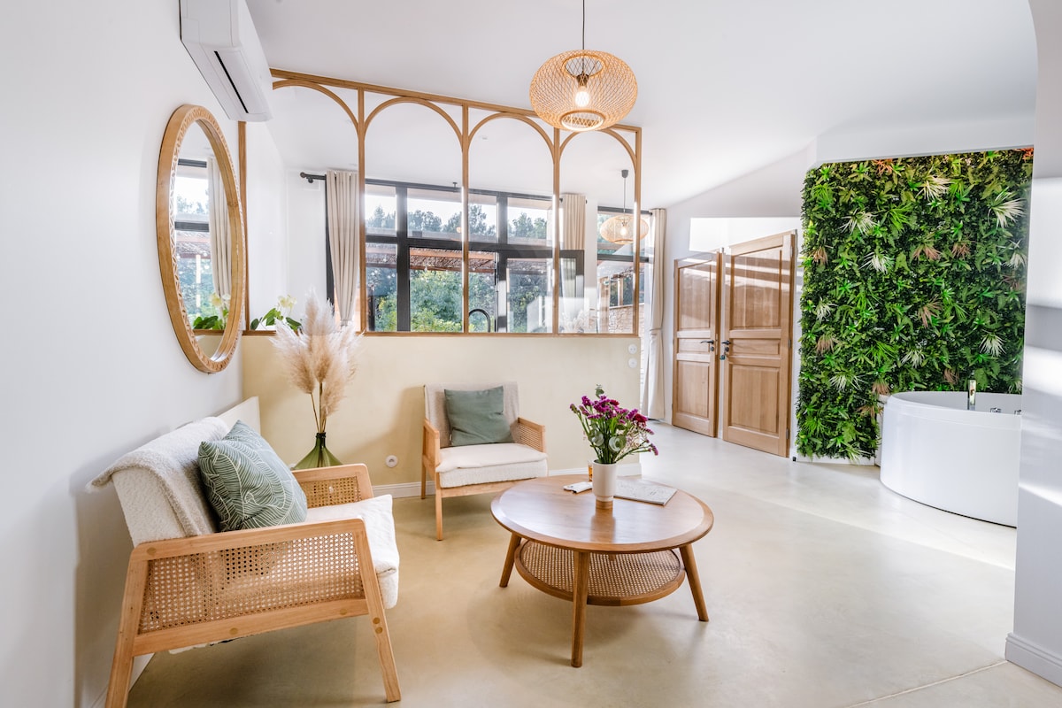 A bright, airy space features a comfortable seating area with natural wood accents. A round table sits at the center, surrounded by light-colored chairs. A lush green wall adds a touch of nature, while large windows provide ample natural light, enhancing the calming atmosphere.