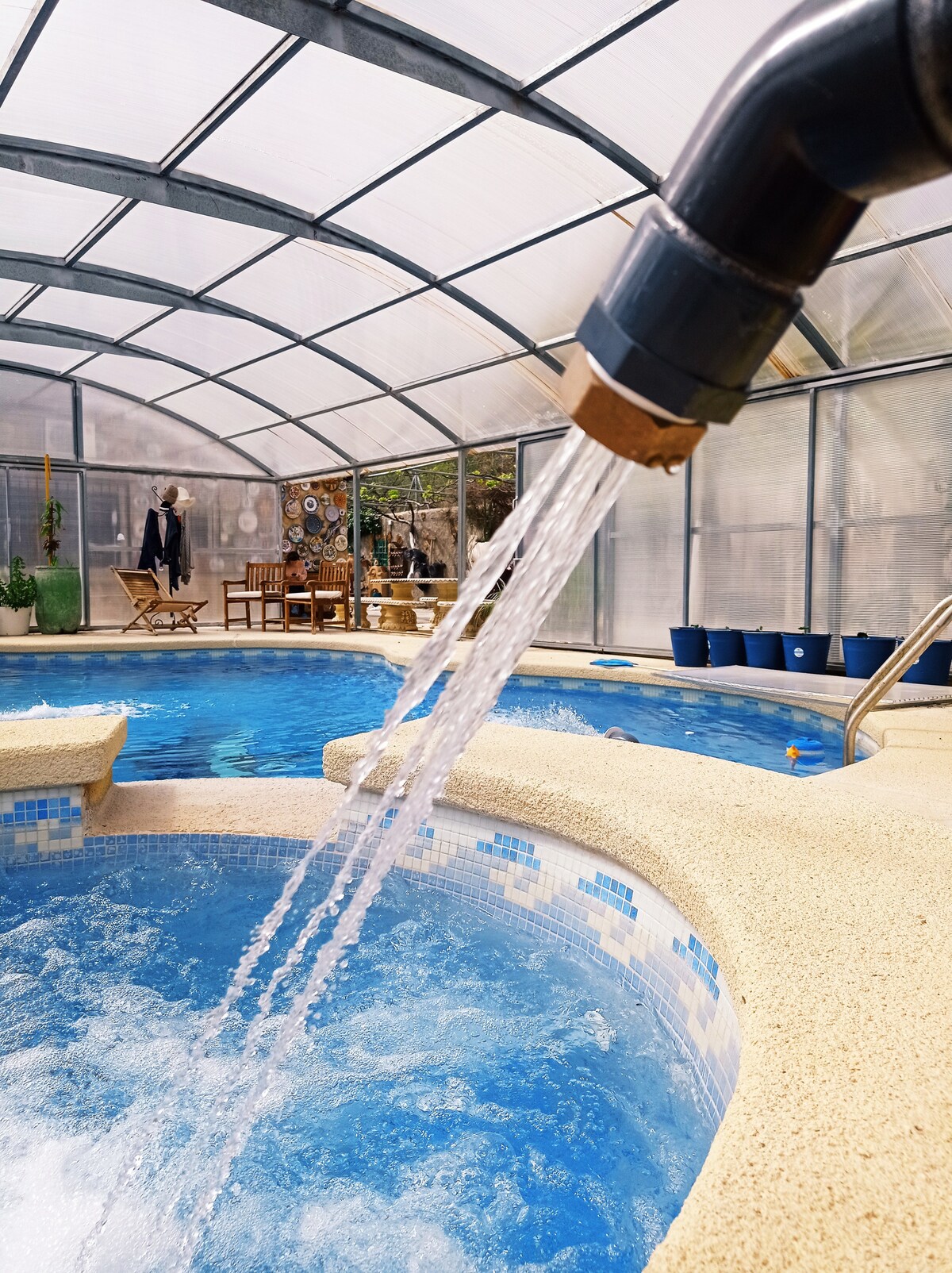 An indoor area features a covered swimming pool with a jacuzzi, both filled with clear blue water. Water flows from a faucet, cascading into the pool. Surrounding the pool, there are lounge chairs and decorative elements enhancing the tranquil setting.