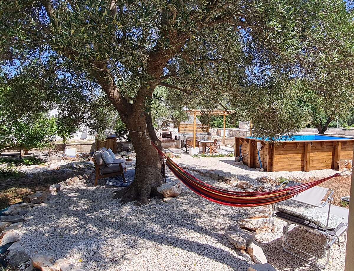 A shaded outdoor area is shown, featuring a hammock suspended between an olive tree and a wooden frame. Comfortable seating arrangements are visible nearby, with a pool nestled in the background, surrounded by rustic wooden decking and stone pathways, creating a serene retreat atmosphere.