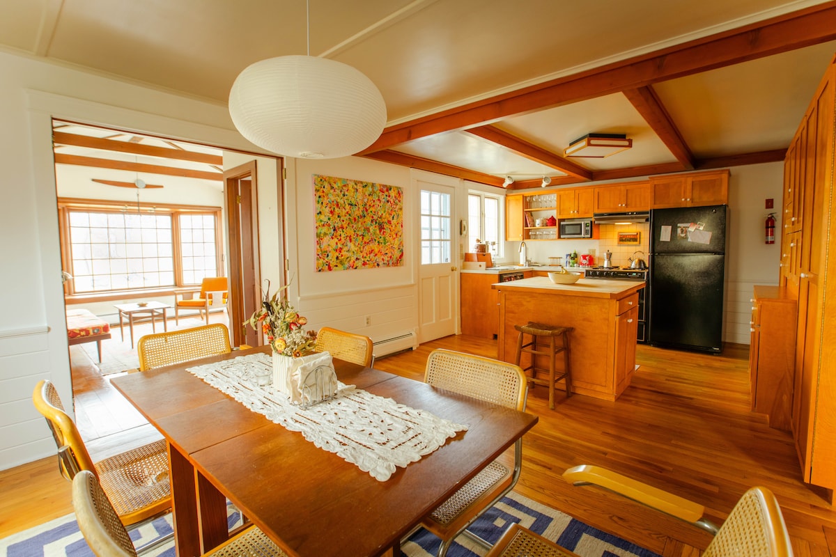 A dining area features a wooden table surrounded by rattan chairs, adorned with a decorative centerpiece. The kitchen is visible in the background, equipped with modern appliances and custom wood cabinetry. Natural light filters through large windows, enhancing the warm hues of the wooden furnishings.