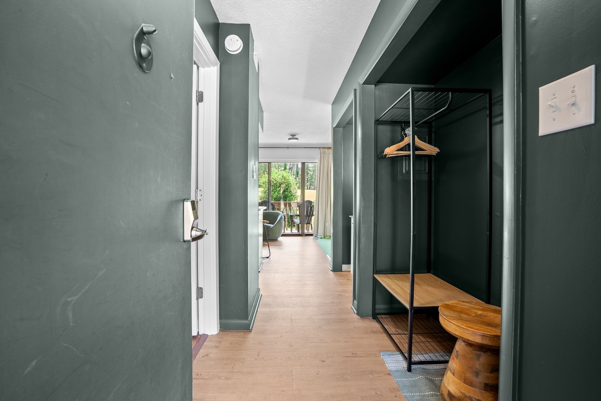A modern entrance foyer is presented with a wardrobe area for hanging storage. A wood bench with a round table provides additional space. Natural light is visible through the doorway leading to an outdoor patio, enhancing the inviting layout.