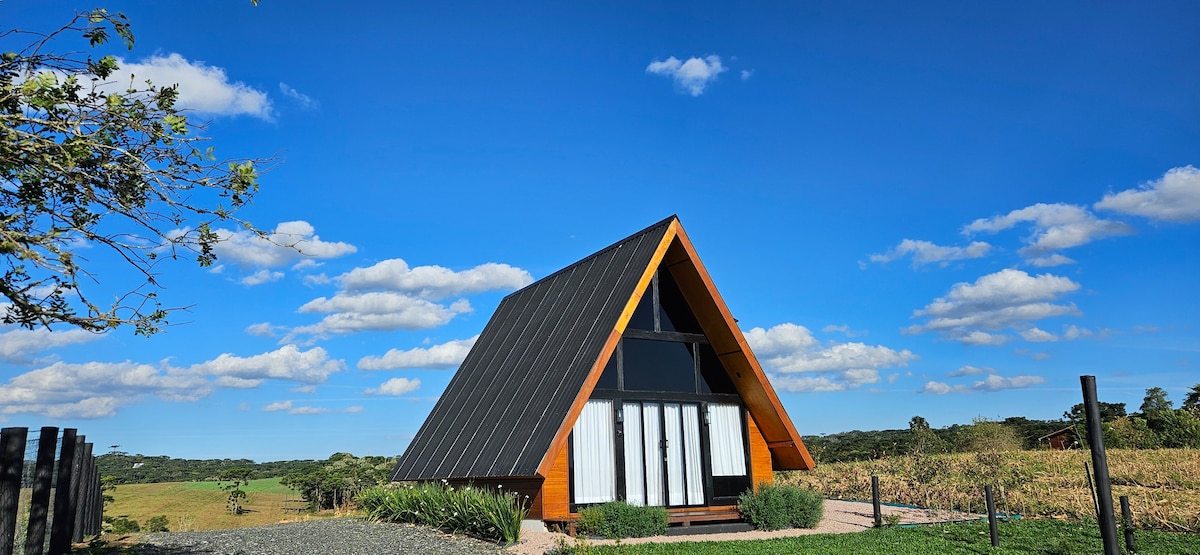 A stylish A-frame cabin is set against a backdrop of clear blue skies and gentle clouds. The structure features large glass doors that invite natural light and provide views of the surrounding countryside.