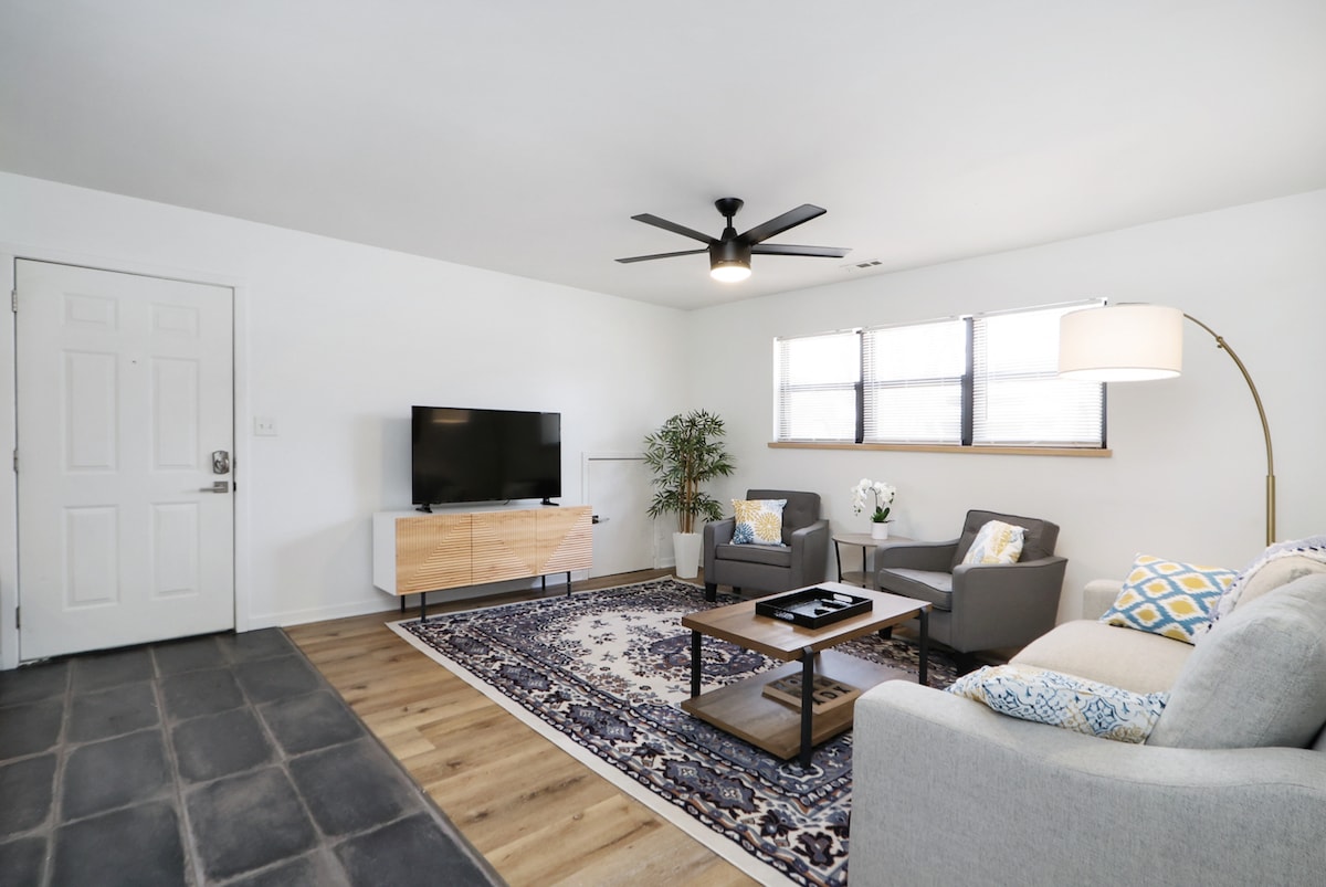 A bright and airy living room features a ceiling fan and a flat-screen television mounted on the wall. Two comfortable armchairs and a sofa are arranged around a wooden coffee table, with a decorative rug adding warmth to the space. Natural light enters through two windows.
