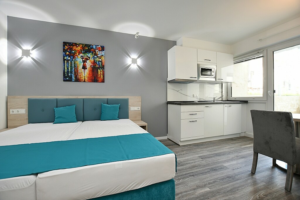 A modern hotel room features a spacious bed with a light blue accent blanket. It includes a kitchenette with a microwave and a dining area with a gray chair. Natural light floods the space through sliding glass doors, which open to a private outdoor area.