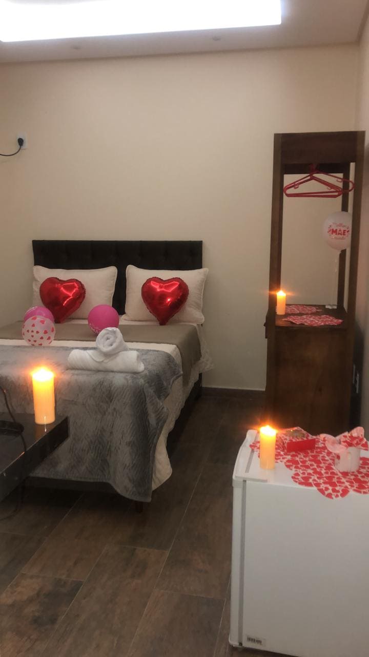 A cozy sleeping area features a spacious bed with soft bedding and two heart-shaped balloons. A small table holds decorative items, while soft candlelight adds a warm ambiance. A mini-fridge and a wooden wardrobe are positioned in the room, offering practical amenities.
