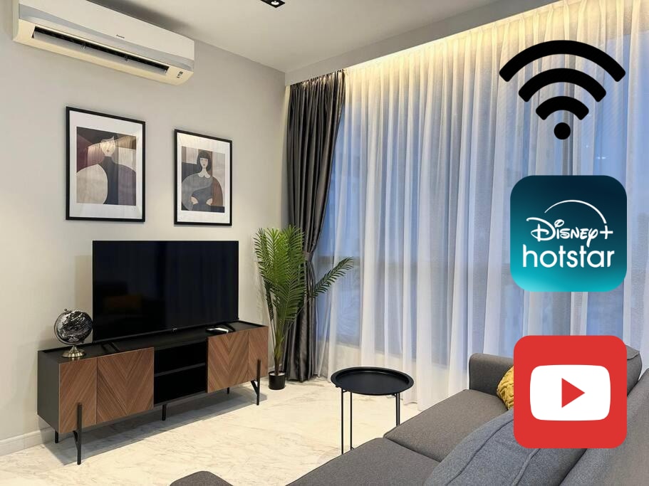 A modern living area is presented with a large Smart TV positioned on a sleek wooden cabinet. Framed artwork decorates the walls, and a potted plant adds a touch of greenery. Natural light filters through sheer curtains, enhancing the inviting space.