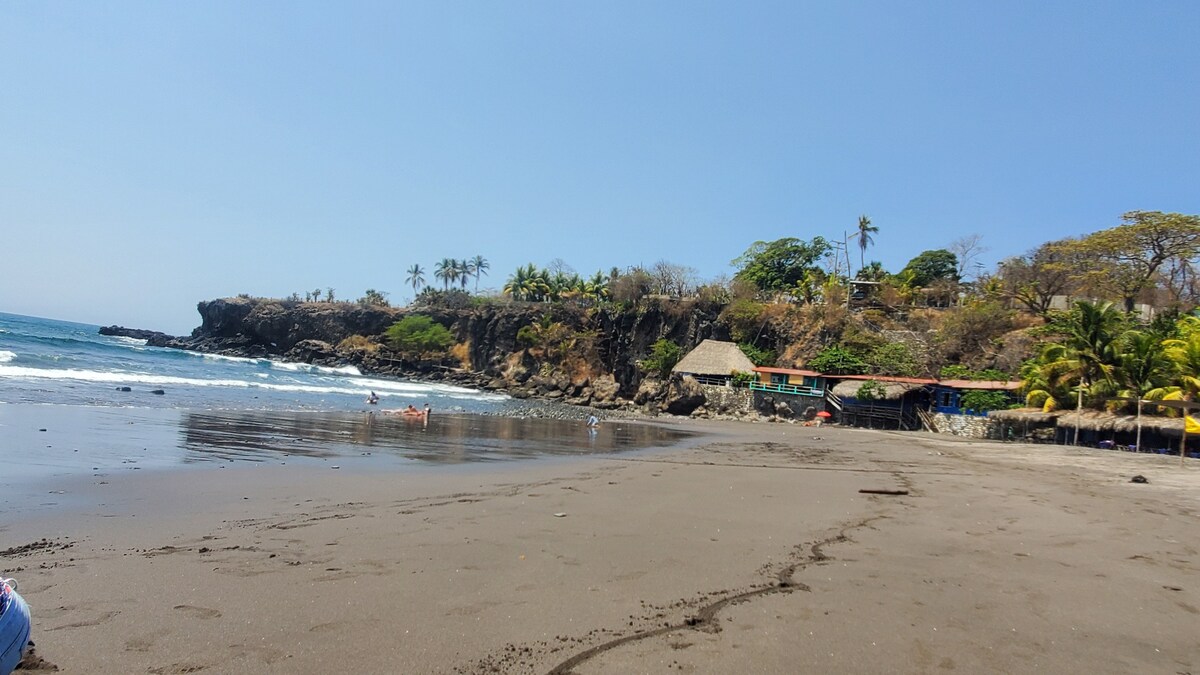 An inviting beach scene features golden sand stretching towards the shimmering blue ocean. Lush palm trees line the coast, and rocky cliffs are visible in the background. Various structures, including a thatched-roof hut, are nestled among greenery, offering shade and a place to relax.