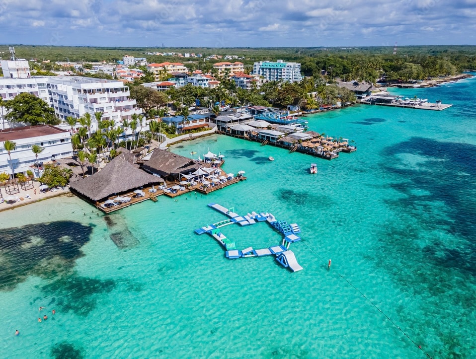 An aerial view captures a vibrant coastal scene, with clear turquoise waters and floating platforms amidst a lively beach atmosphere. Various boats are moored near the shoreline, while beachfront establishments and lush greenery are visible in the background, enhancing the inviting coastal environment.