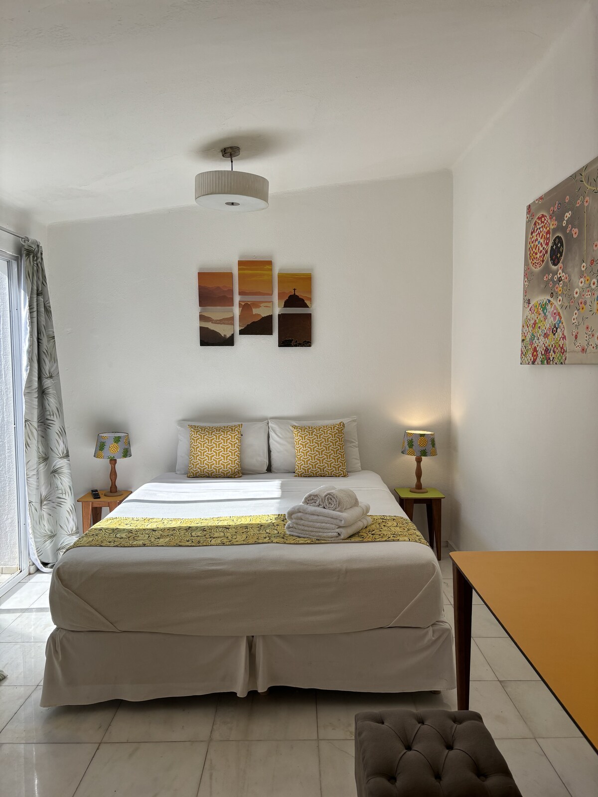 A well-lit sleeping area features a spacious bed with decorative pillows and a light blanket. Two bedside tables hold stylish lamps, and a colorful wall art piece adds character. Natural light enters through glass doors leading to a small outdoor area.