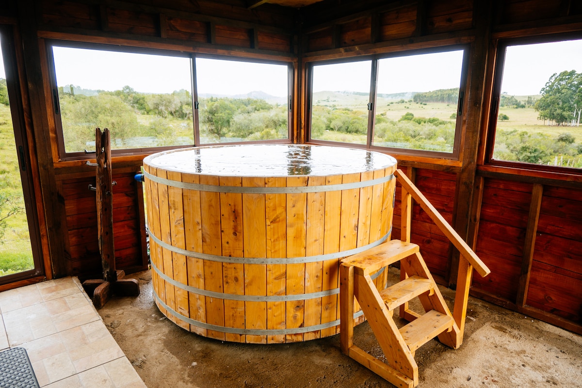 A wooden hot tub is placed in a spacious area surrounded by large windows, offering views of the natural landscape. Sturdy wooden steps lead up to the tub, enhancing accessibility. The warm wood tones complement the earthy surroundings, creating a serene atmosphere.
